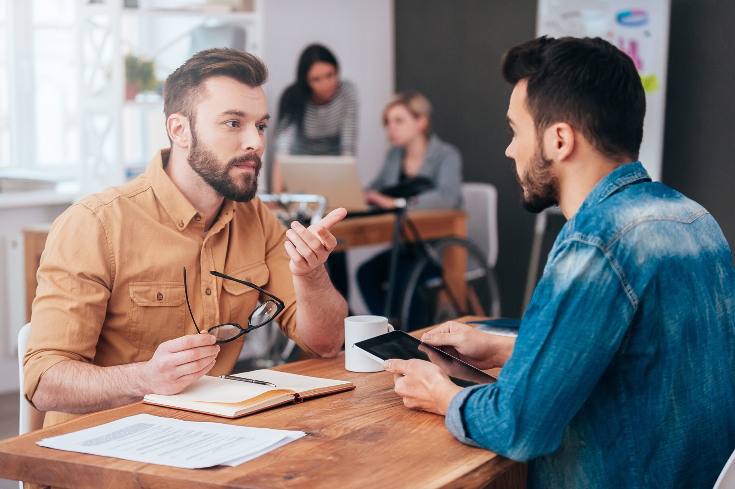 Two confident young men talking and gesturing while sitting at the desk in office with two colleagues